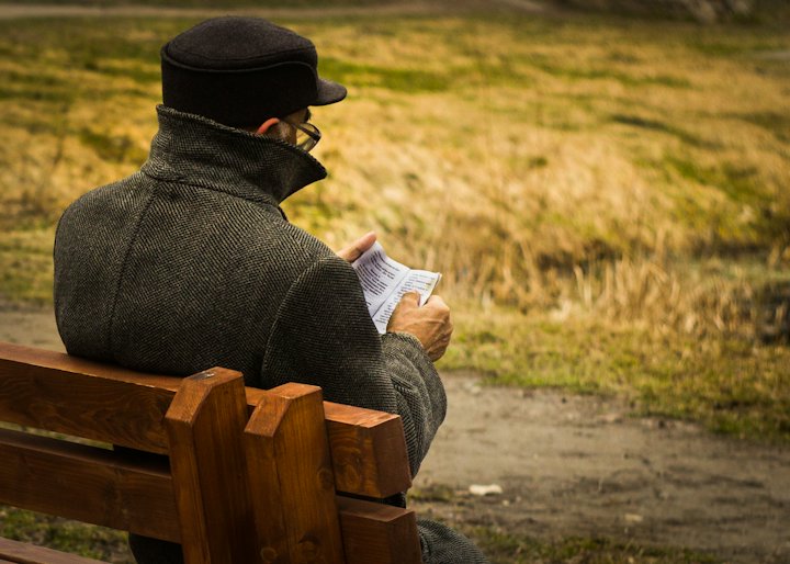 A man in a cap and coat sitting on a wooden bench, reading a sheet of paper in the afternoon sun
