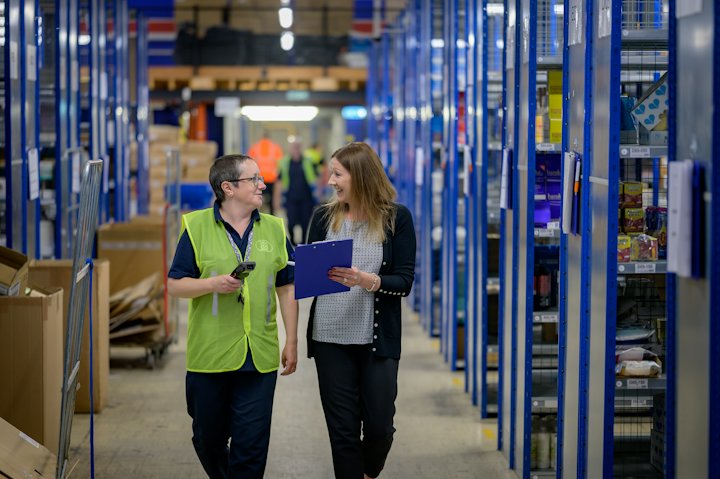 Two women in high-visibility vests walking through a warehouse aisle, one holding a folder
