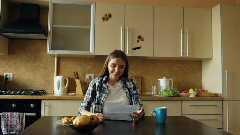 A woman reading a letter at her kitchen table, with a mug and a bowl of fruit in front of her